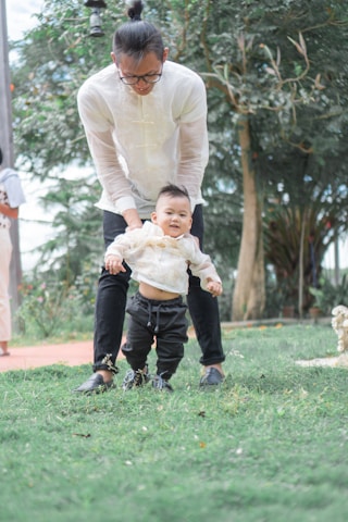 An adult is gently guiding a baby as they practice walking on grass. The scene is set outdoors with trees in the background, giving a serene and natural environment. The adult is wearing glasses and a light-colored top, while the baby is dressed in casual clothes.