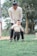 An adult is gently guiding a baby as they practice walking on grass. The scene is set outdoors with trees in the background, giving a serene and natural environment. The adult is wearing glasses and a light-colored top, while the baby is dressed in casual clothes.