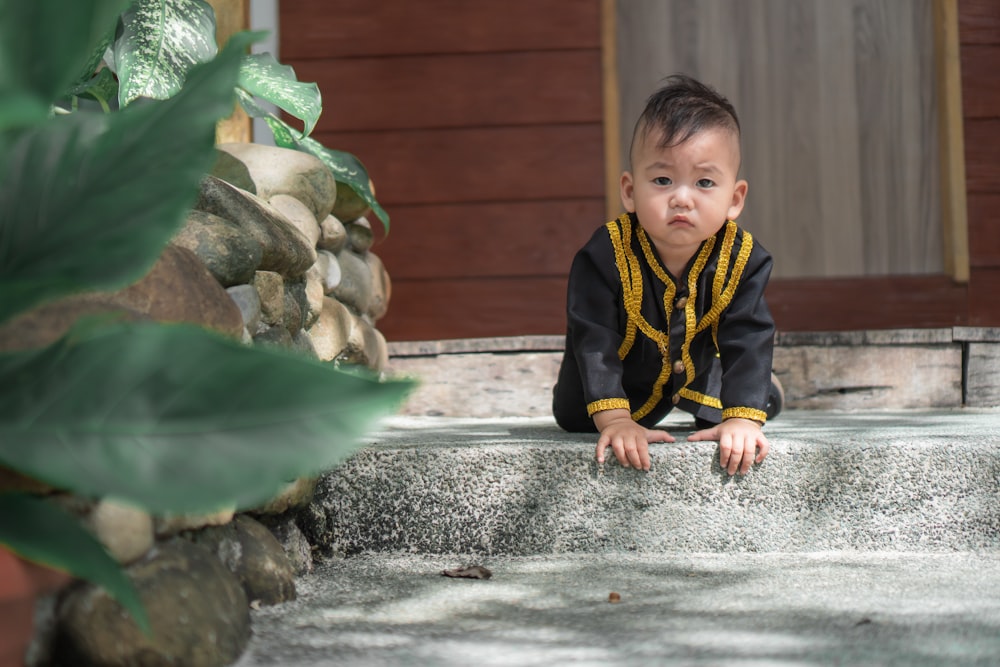 boy in black and yellow zip up jacket sitting on gray concrete fountain during daytime
