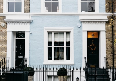 A charming facade of a townhouse featuring a light blue brick exterior with white trim. The central area includes a large window with multiple panes, flanked by two black doors each adorned with decorative elements. A black wrought iron fence runs along the front, with topiary shrubs in containers placed symmetrically. Two security alarms are visible on the upper part of the facade, and a decorative wreath hangs on one of the doors.
