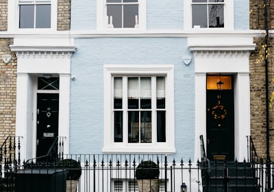 A charming facade of a townhouse featuring a light blue brick exterior with white trim. The central area includes a large window with multiple panes, flanked by two black doors each adorned with decorative elements. A black wrought iron fence runs along the front, with topiary shrubs in containers placed symmetrically. Two security alarms are visible on the upper part of the facade, and a decorative wreath hangs on one of the doors.