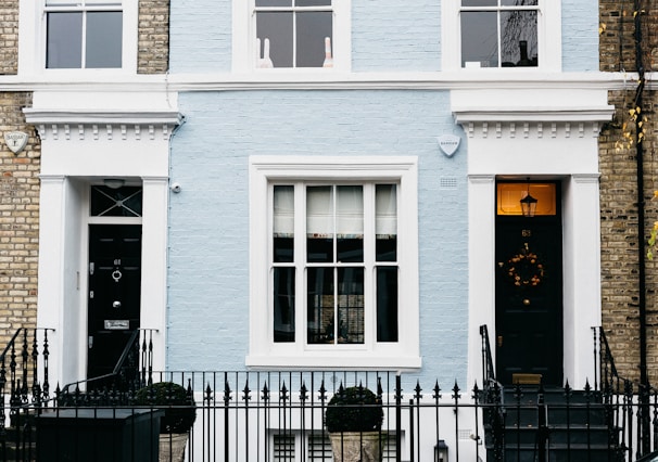 A charming facade of a townhouse featuring a light blue brick exterior with white trim. The central area includes a large window with multiple panes, flanked by two black doors each adorned with decorative elements. A black wrought iron fence runs along the front, with topiary shrubs in containers placed symmetrically. Two security alarms are visible on the upper part of the facade, and a decorative wreath hangs on one of the doors.