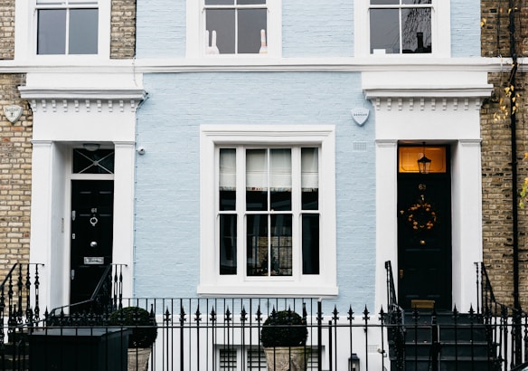 A charming facade of a townhouse featuring a light blue brick exterior with white trim. The central area includes a large window with multiple panes, flanked by two black doors each adorned with decorative elements. A black wrought iron fence runs along the front, with topiary shrubs in containers placed symmetrically. Two security alarms are visible on the upper part of the facade, and a decorative wreath hangs on one of the doors.
