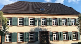 A two-story building featuring a beige facade with green shutters on all the windows. The roof is dark-colored and sloped, with a few small skylights. The name 'St. Josef's Heim' is visible above the main entrance. Some trees are partially visible on either side of the building.