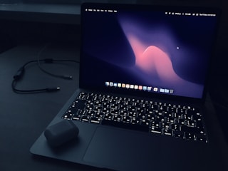 A laptop with an illuminated keyboard sits on a dark surface. The screen displays a dark abstract wallpaper with a wave-like pattern in shades of pink and purple. An open case for wireless earbuds is placed on the laptop's wrist rest. In the background, a charging cable is visible.
