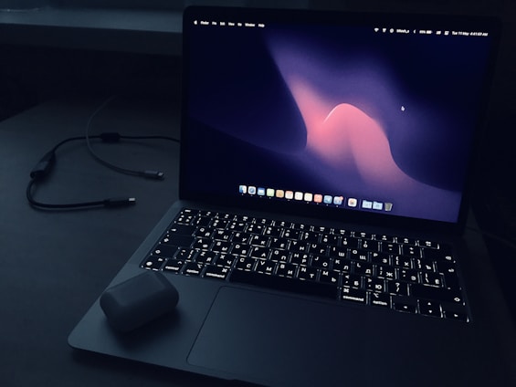 A laptop with an illuminated keyboard sits on a dark surface. The screen displays a dark abstract wallpaper with a wave-like pattern in shades of pink and purple. An open case for wireless earbuds is placed on the laptop's wrist rest. In the background, a charging cable is visible.