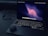 A laptop with an illuminated keyboard sits on a dark surface. The screen displays a dark abstract wallpaper with a wave-like pattern in shades of pink and purple. An open case for wireless earbuds is placed on the laptop's wrist rest. In the background, a charging cable is visible.