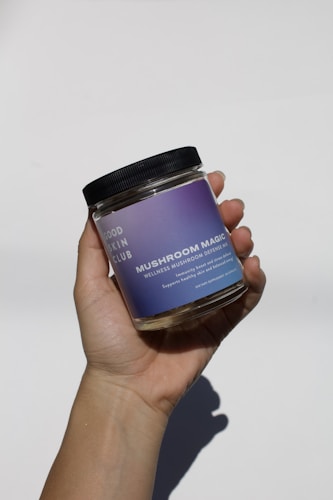 A hand is holding a glass jar with a black lid. The jar has a purple gradient label with the text 'GOOD SKIN CLUB' and 'MUSHROOM MAGIC WELLNESS MUSHROOM DEFENSE MIX' along with additional information about its benefits.