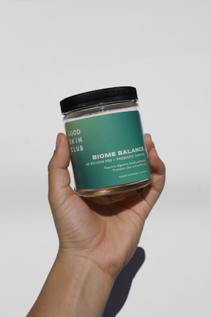 A hand is holding a clear jar with a green label that reads 'GOOD SKIN CLUB BIOME BALANCE 40 BILLION PRE + PROBIOTIC SUPPORT. Supports digestive health and balance. Promotes clear and healthy skin. DIETARY SUPPLEMENT IN CAPSULES.' The jar has a black screw-top lid.