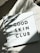 A white box with the text 'GOOD SKIN CLUB' printed in bold black letters sits on a light-colored surface. Shadows of leaves are cast on the box, giving it a natural and serene appearance.