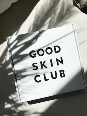 A white box with the text 'GOOD SKIN CLUB' printed in bold black letters sits on a light-colored surface. Shadows of leaves are cast on the box, giving it a natural and serene appearance.