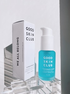 A skincare product consisting of a white rectangular box and a blue serum bottle displayed on a clear acrylic stand. The box has bold black text saying 'Good Skin Club' and 'We All Belong'. The bottle is labeled as 'Ultrahydrate Serum' and contains essential amino acids.