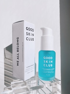 A skincare product consisting of a white rectangular box and a blue serum bottle displayed on a clear acrylic stand. The box has bold black text saying 'Good Skin Club' and 'We All Belong'. The bottle is labeled as 'Ultrahydrate Serum' and contains essential amino acids.