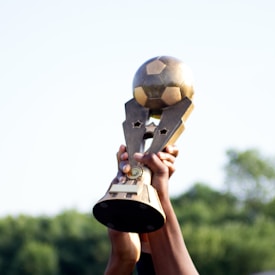 A trophy shaped like a soccer ball being lifted in the air by multiple hands, set against a blurred background of trees and a clear sky.