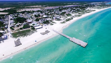 Aerial view of a coastal town with a white sandy beach, stretching into clear turquoise waters. The beach features several shaded umbrellas and a long pier extending into the ocean. A dense cluster of buildings and green vegetation is visible inland.