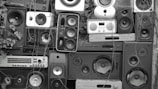 A collage of various speakers and sound equipment stacked together. The arrangement includes different sizes and styles of speakers, some with visible wear and tear. Overgrown plants and vines are interspersed among the equipment, creating a contrast between technology and nature.
