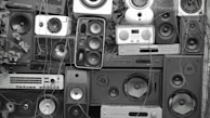 A collage of various speakers and sound equipment stacked together. The arrangement includes different sizes and styles of speakers, some with visible wear and tear. Overgrown plants and vines are interspersed among the equipment, creating a contrast between technology and nature.