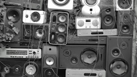 A collage of various speakers and sound equipment stacked together. The arrangement includes different sizes and styles of speakers, some with visible wear and tear. Overgrown plants and vines are interspersed among the equipment, creating a contrast between technology and nature.