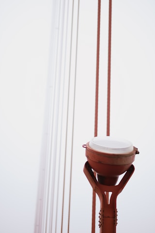 A close-up view of an orange structural element that appears to be part of a large bridge, with thick cables leading upwards into the foggy sky.