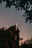 A brick church with a prominent illuminated cross atop its roof, surrounded by silhouetted trees against a dusky sky.