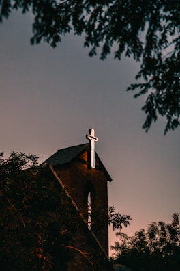 A brick church with a prominent illuminated cross atop its roof, surrounded by silhouetted trees against a dusky sky.