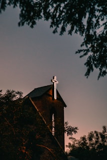 A brick church with a prominent illuminated cross atop its roof, surrounded by silhouetted trees against a dusky sky.