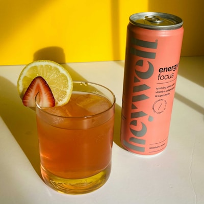 A glass of orange-colored beverage with ice, garnished with a lemon slice and a strawberry wedge, is placed beside a tall, pink can of energy drink. The background is a bright yellow, creating a vibrant contrast.