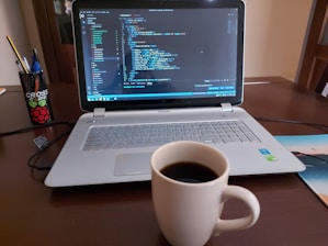 A laptop displaying code on the screen with a notepad and coffee cup beside it.