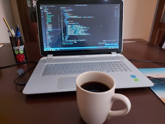 A laptop displaying code is placed on a wooden table next to a white mug filled with coffee. To the left, there is a black holder with pens and pencils. The screen shows a coding interface with various colors denoting syntax highlighting.