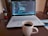 A laptop displaying code is placed on a wooden table next to a white mug filled with coffee. To the left, there is a black holder with pens and pencils. The screen shows a coding interface with various colors denoting syntax highlighting.