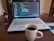 A laptop displaying code is placed on a wooden table next to a white mug filled with coffee. To the left, there is a black holder with pens and pencils. The screen shows a coding interface with various colors denoting syntax highlighting.