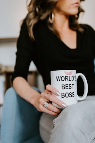 A woman is sitting on a blue chair holding a white mug with the text 'WORLD'S BEST BOSS' and a lipstick mark on it. She is wearing a black top and grey jeans. Her hand is adorned with rings and has silver nail polish.