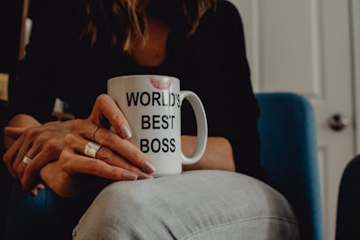 A person is sitting and holding a white mug with the text 'WORLD'S BEST BOSS' printed on it. The mug has a lipstick mark near the rim. The person is wearing rings on their fingers and is dressed in casual attire. The background includes a blue chair and a partially visible door.