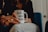 A person is sitting and holding a white mug with the text 'WORLD'S BEST BOSS' printed on it. The mug has a lipstick mark near the rim. The person is wearing rings on their fingers and is dressed in casual attire. The background includes a blue chair and a partially visible door.