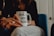 A person is sitting and holding a white mug with the text 'WORLD'S BEST BOSS' printed on it. The mug has a lipstick mark near the rim. The person is wearing rings on their fingers and is dressed in casual attire. The background includes a blue chair and a partially visible door.