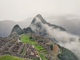An ancient stone city is set on a mountainside surrounded by lush green terraces and steep cliffs. Mist and clouds hover around, partially obscuring the view of distant mountains. The stone structures display intricate architecture and pathways, offering a glimpse into a historical civilization.