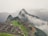 An ancient stone city is set on a mountainside surrounded by lush green terraces and steep cliffs. Mist and clouds hover around, partially obscuring the view of distant mountains. The stone structures display intricate architecture and pathways, offering a glimpse into a historical civilization.
