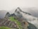 An ancient stone city is set on a mountainside surrounded by lush green terraces and steep cliffs. Mist and clouds hover around, partially obscuring the view of distant mountains. The stone structures display intricate architecture and pathways, offering a glimpse into a historical civilization.