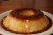 A caramel custard dessert sits on a white plate. The flan is smooth and glossy, with a rich caramel sauce cascading down its sides, creating a glistening appearance. The setting has a warm ambiance with a wooden surface in the background.