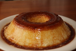 A caramel custard dessert sits on a white plate. The flan is smooth and glossy, with a rich caramel sauce cascading down its sides, creating a glistening appearance. The setting has a warm ambiance with a wooden surface in the background.