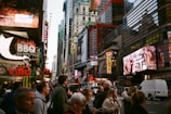 A bustling urban street scene with a crowd of people crossing a street in a busy city area. Numerous brightly lit advertisements and billboards are visible on buildings, showcasing establishments like Dallas BBQ and Villa, alongside various other ads. The scene includes a double-decker bus and vehicles, with a tall mix of historical and modern architecture.