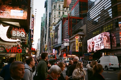 A bustling urban street scene with a crowd of people crossing a street in a busy city area. Numerous brightly lit advertisements and billboards are visible on buildings, showcasing establishments like Dallas BBQ and Villa, alongside various other ads. The scene includes a double-decker bus and vehicles, with a tall mix of historical and modern architecture.