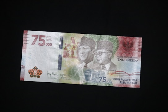 An Indonesian 75,000 Rupiah commemorative banknote featuring images of two prominent figures, national symbols, and a detailed background design. The note shows varied text, intricate patterns, and vibrant colors.