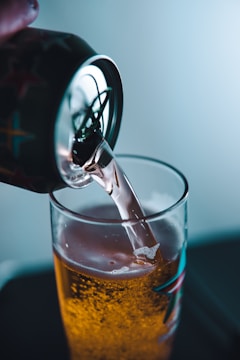 A can is tilted to pour a carbonated beverage into a glass, filling it halfway. The liquid has a golden hue with visible bubbles, and the glass features a decorative pattern.