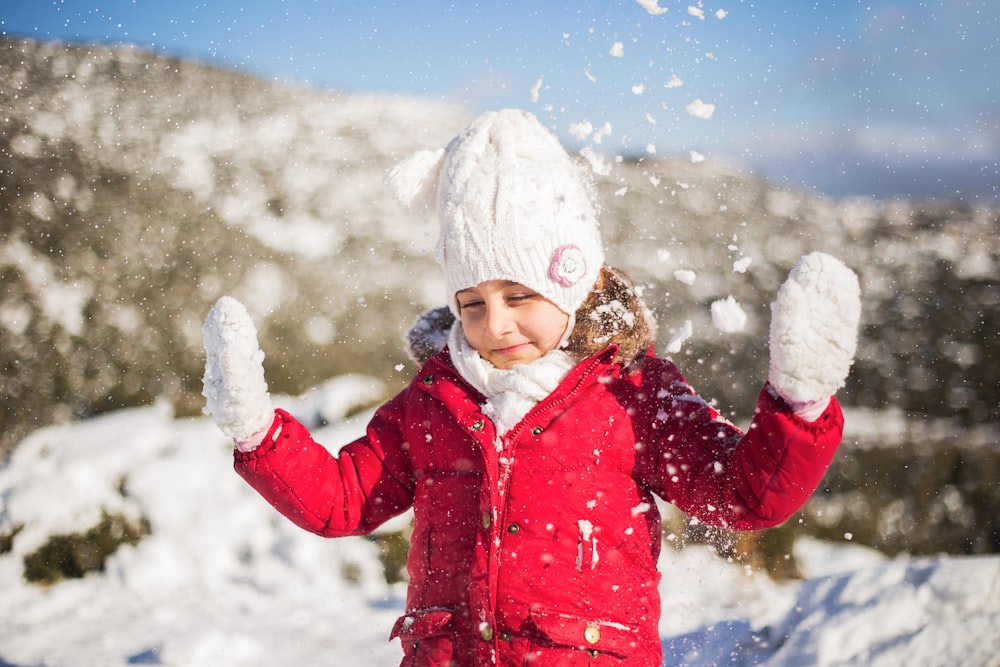 girl in red jacket and white knit cap standing on snow covered ground during daytime