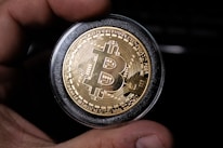 A close-up view of a hand holding a gold-colored coin with the Bitcoin logo prominently displayed in the center. The coin is encased in a protective transparent cover, and intricate circuit-like patterns are etched around the logo, symbolizing digital technology.