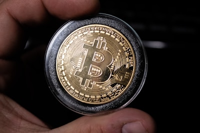 A close-up view of a hand holding a gold-colored coin with the Bitcoin logo prominently displayed in the center. The coin is encased in a protective transparent cover, and intricate circuit-like patterns are etched around the logo, symbolizing digital technology.
