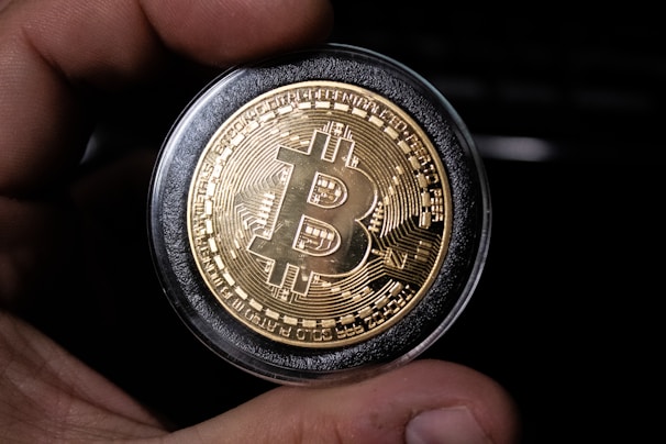 A close-up view of a hand holding a gold-colored coin with the Bitcoin logo prominently displayed in the center. The coin is encased in a protective transparent cover, and intricate circuit-like patterns are etched around the logo, symbolizing digital technology.
