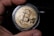 A close-up view of a hand holding a gold-colored coin with the Bitcoin logo prominently displayed in the center. The coin is encased in a protective transparent cover, and intricate circuit-like patterns are etched around the logo, symbolizing digital technology.