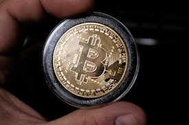 A close-up view of a hand holding a gold-colored coin with the Bitcoin logo prominently displayed in the center. The coin is encased in a protective transparent cover, and intricate circuit-like patterns are etched around the logo, symbolizing digital technology.